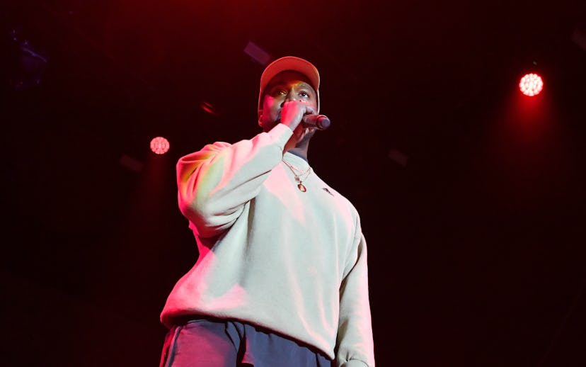 Kanye West on stage in a white sweatshirt and a white cap with a microphone in his hands