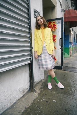 A brunette woman in a yellow shirt, yellow blazer, orange tie, grey check skirt, green socks and pin...