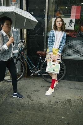 A brunette woman in a blue checked, white and yellow checked dress, red socks, beige shoes and bag a...