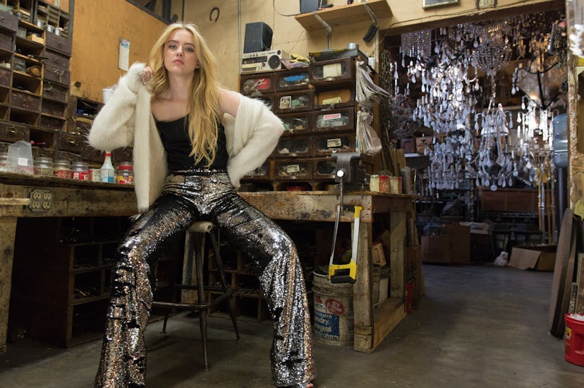 Kathryn Newton in a black shirt, shiny pants and a white cardigan sitting on a bar chair 