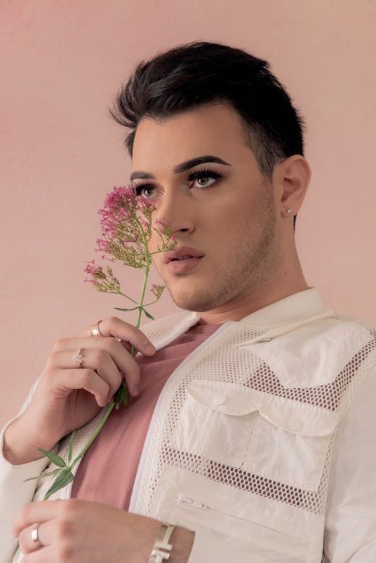 Manny Mua, in a pink shirt and white jacket, holding a light pink flower next to his face
