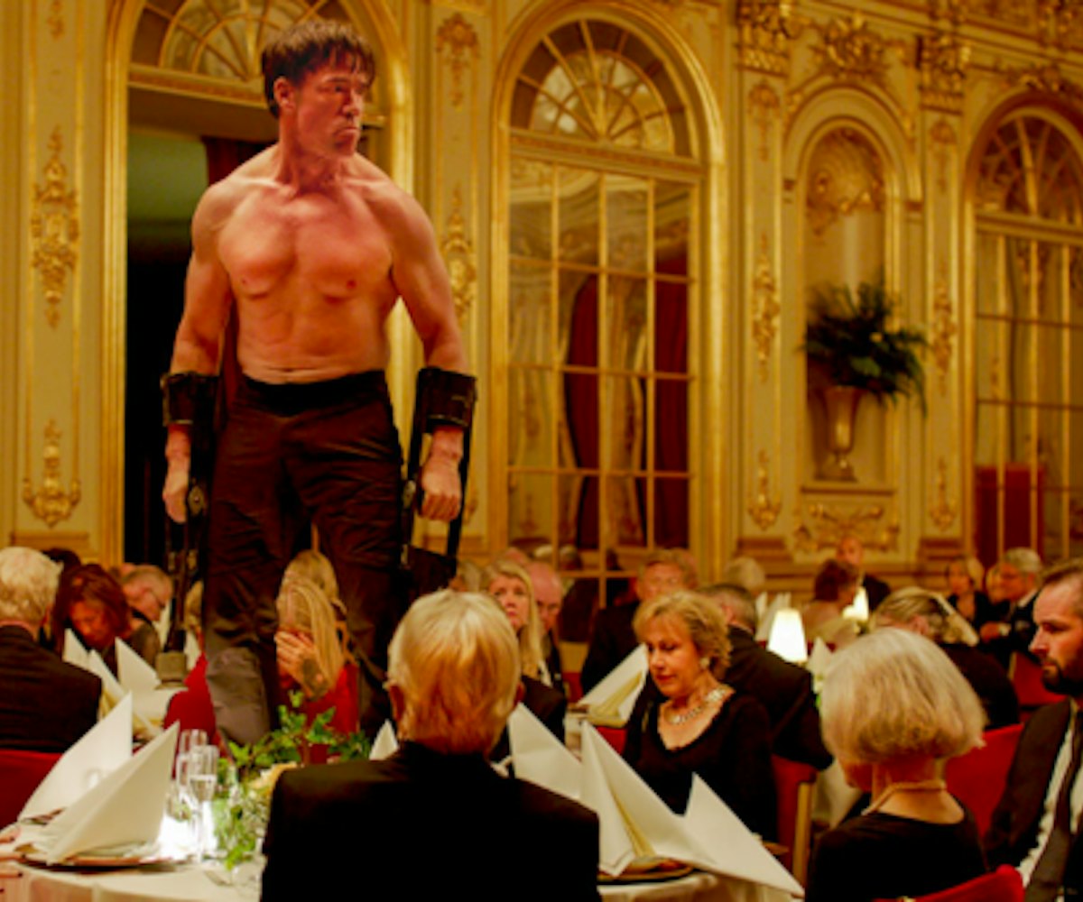 Actor Terry Notary wearing no shirt, standing on a table with people surrounding him and dining