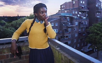 Michaela Coel standing on a balcony in a yellow sweater and dark blue skirt in a scene from Chewing ...