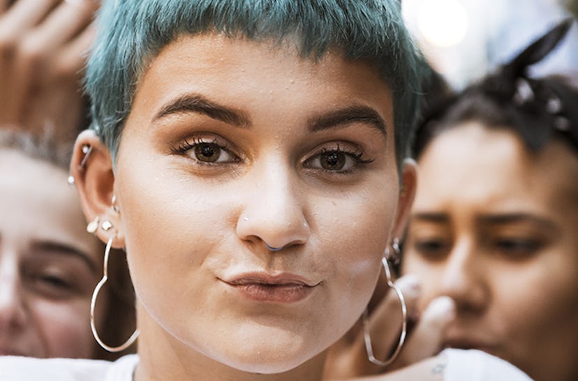 A woman with clumped eyelashes, blue hair and large heart-shaped earrings pouting at the camera 