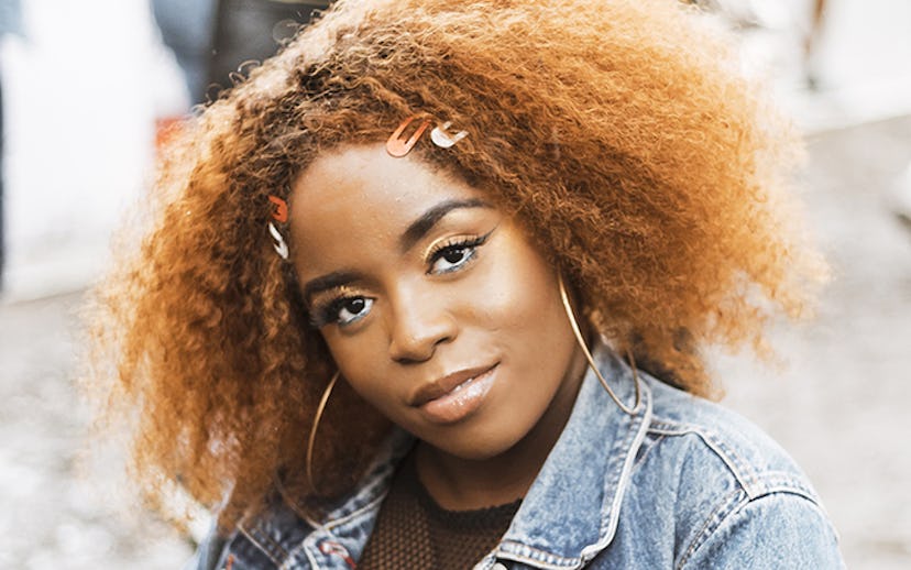 A woman with afro hair, graphic eyeliner, wearing a denim jacket, smiling