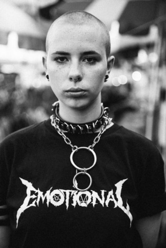 A bald young person wearing a huge choker necklace with chains and a black t-shirt with the text "Em...