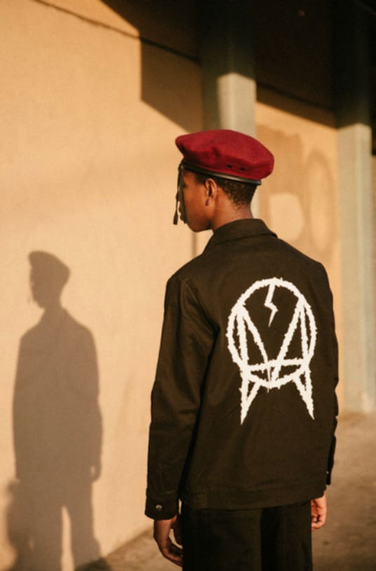 A young man posing with his back turned while wearing a red beret and a black jacket with a white lo...