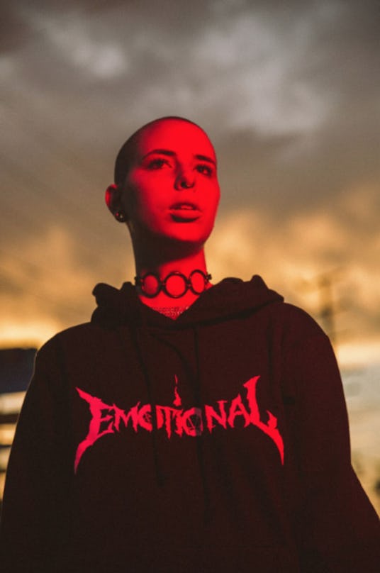 A young bald person standing in red lighting while wearing a choker made of hoops and a black hoodie...