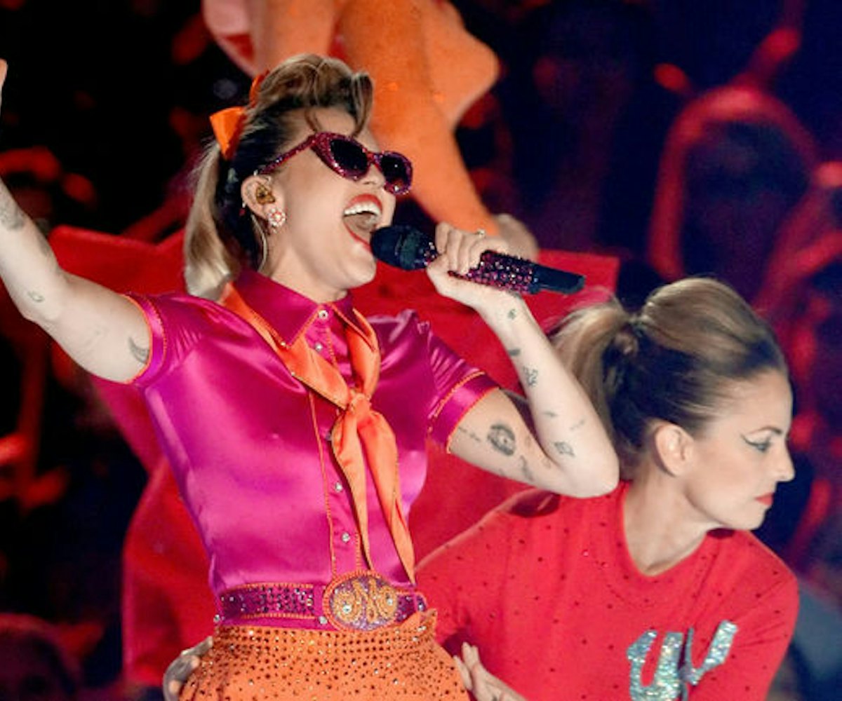 Singer Miley Cyrus wearing a pink shirt and orange skirt and tie, while singing in a mic with a girl...