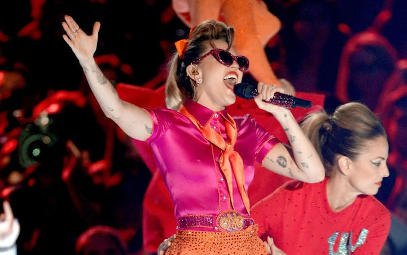 Miley Cyrus wearing a pink shirt and orange skirt and tie, while singing on stage