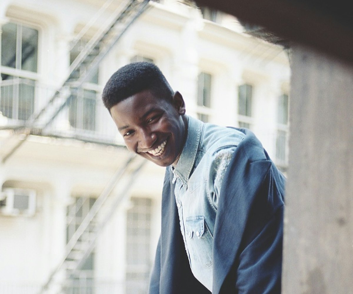 Mamoudou Athie is standing and smiling while leaning against the balcony railing in a blue denim shi...