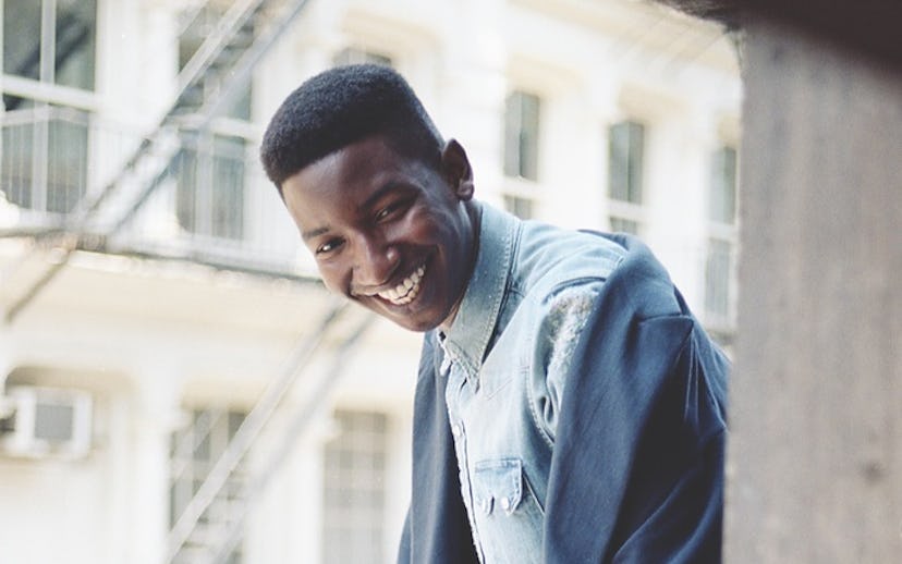 Mamoudou Athie is standing and smiling while leaning against the balcony railing in a blue denim shi...