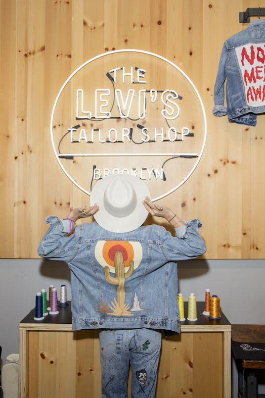 A girl looking up at "The Levi's Tailor Shop" sign in her jean jacket that has a cactus sticker on i...