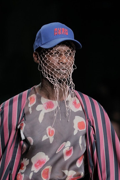 A male model in a printed gray shirt, gray and pink striped silk jacket, baseball hat and a white ne...