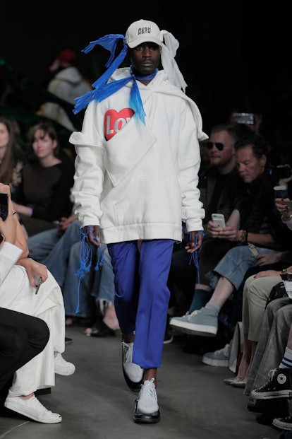 A male model in a white sweatshirt, blue trousers, baseball hat and trash bags under the hat and aro...
