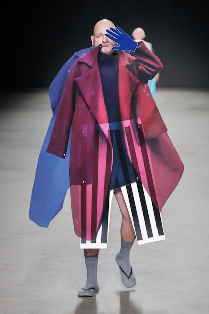 A male model holding plastic clothing items, like striped pants, pink and blue jacket