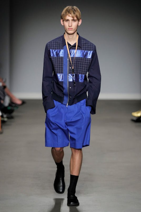 A male model in an all blue outfit, shorts, shirt and jacket with some detail, and black shoes and s...