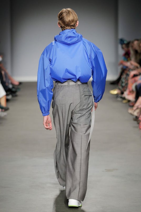 A male model from behind wearing a blue windbreaker jacket and gray trousers