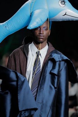 A male model in a suit that has blue and brown details wearing a blow-up dolphin on his head