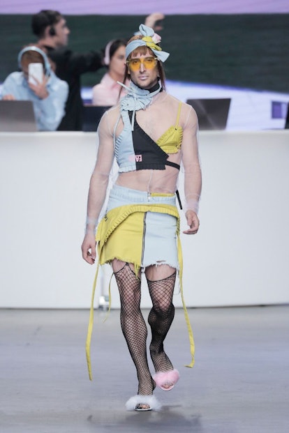 A male model in blue and yellow skirt and a matching top and hairpiece,  fishnet tights and fluffy s...