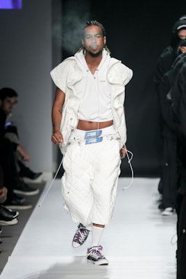 A male model in an all-white outfit that includes pants, cropped sweatshirt and a vest