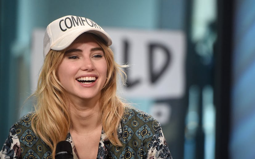 Suki Waterhouse doing an interview while holding a mic and wearing a green silk shirt and white cap