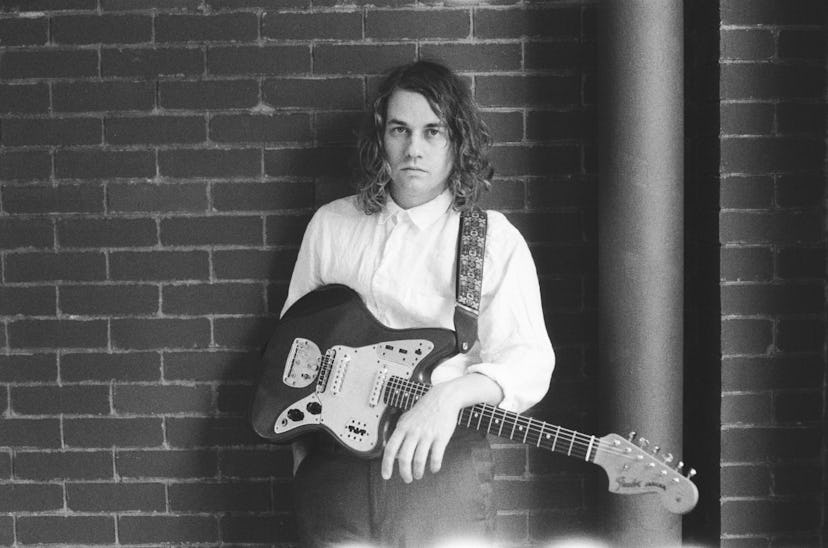 Kevin Morby leaning against a brick wall, holding a guitar in his lap 