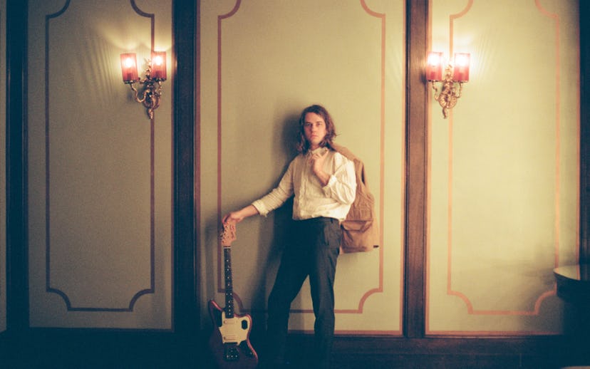 Kevin Morby leaning against a wall, holding a guitar