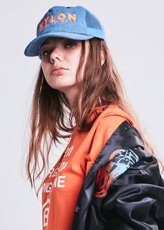 Model posing while wearing a blue cap, an orange shirt, and a black jacket 