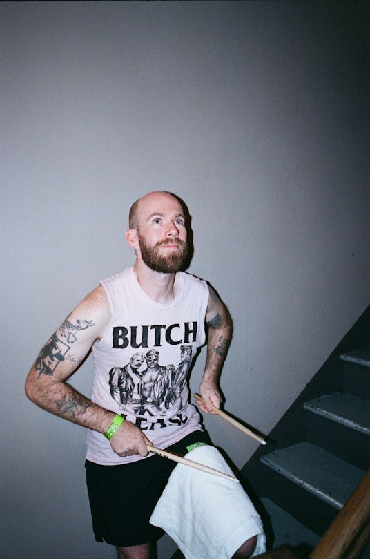 A man with a "Butch Please" shirt and drum sticks leaning against a white wall holding drumsticks