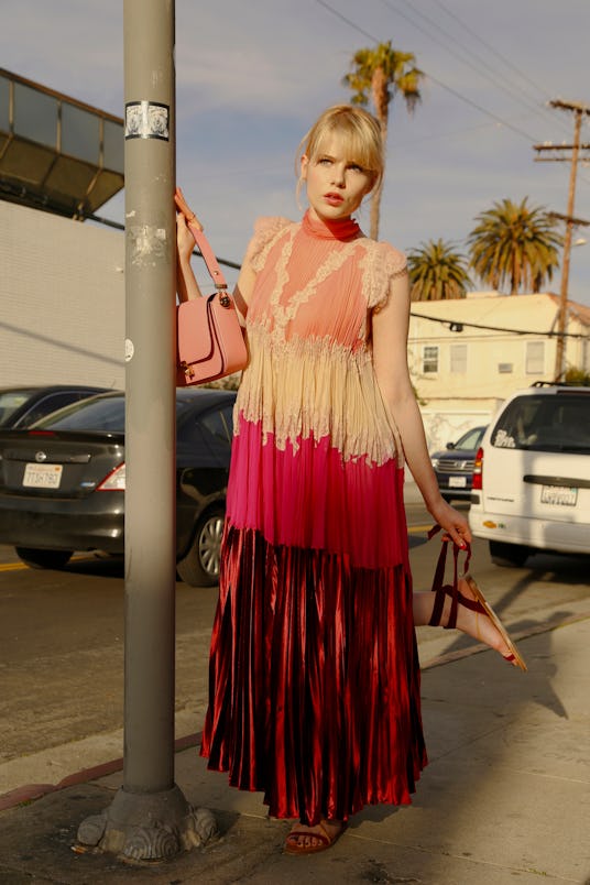 Lucy Boynton in a white and pink dress standing next to a pole in the street in Sing Street