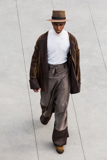A male model in a white shirt, leather jacker and hat walking down the runway