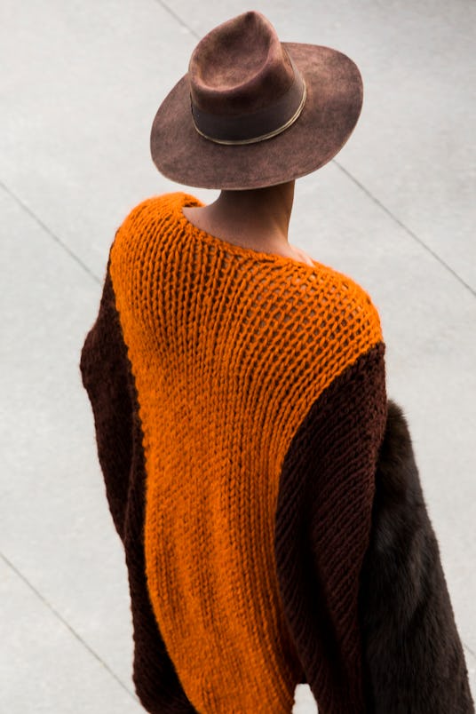A male model in a brown and orange sweater and leather hat walking down the runway