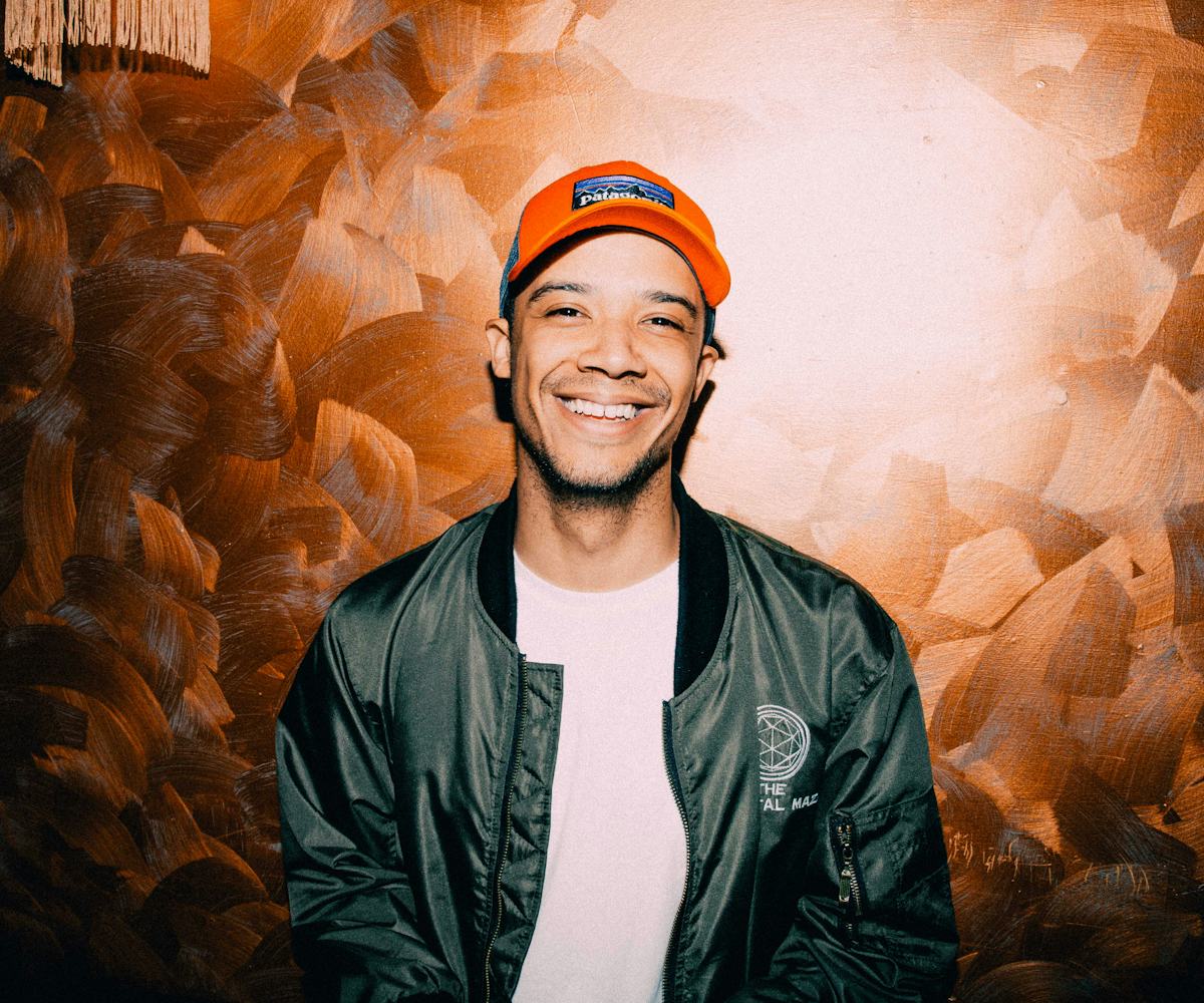 Raleigh Ritchie posing in front of a brown wall while wearing a green jacket and an orange cap