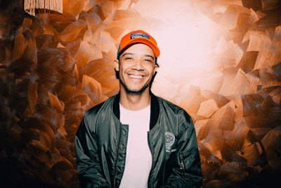 Raleigh Ritchie posing in front of a brown wall, wearing a green jacket and orange cap.