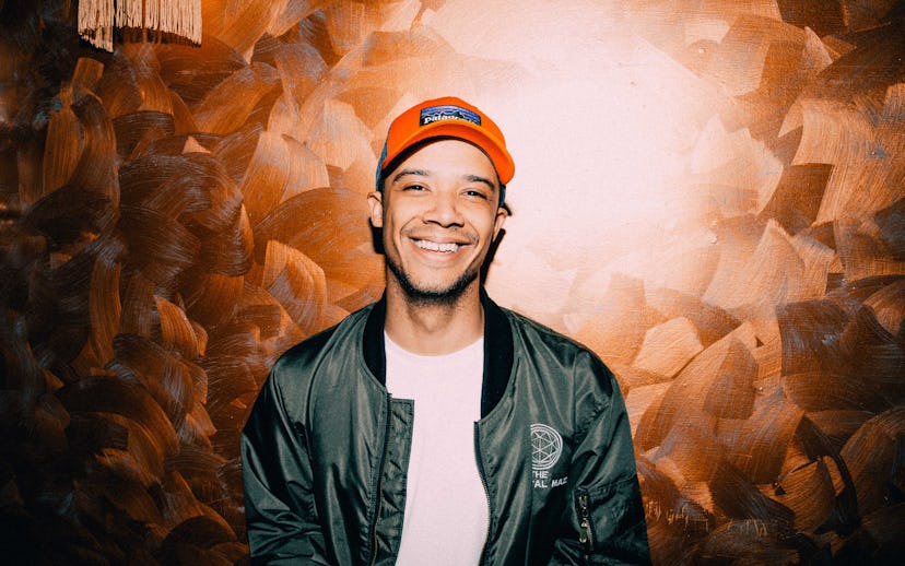 Raleigh Ritchie posing in front of a brown wall while wearing a green jacket and an orange cap
