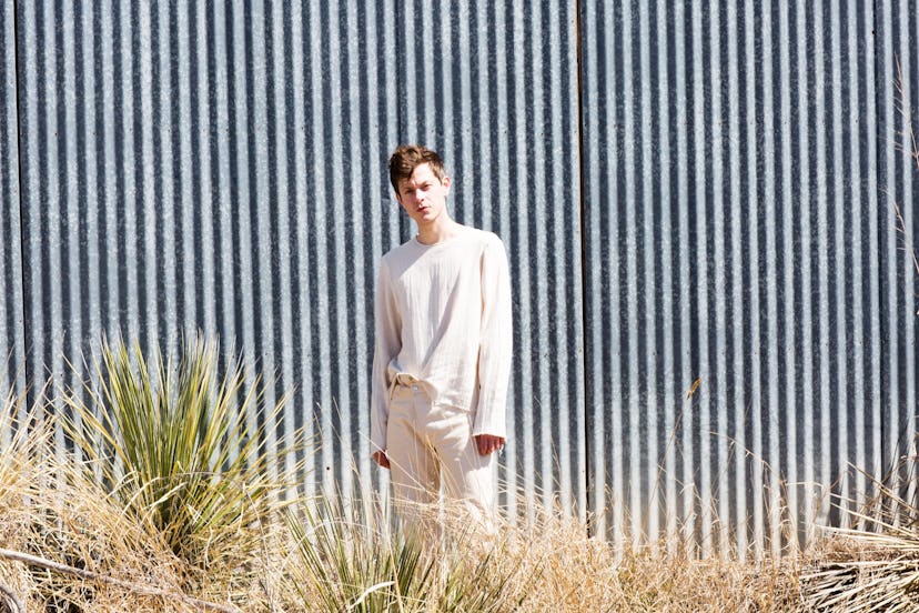 Michael Hadreas in all white standing in front of a striped black and white wall in a desert