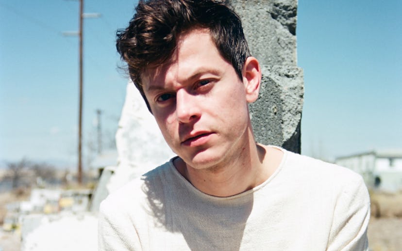 Michael Hadreas in all white crouching in front of a rock in a desert