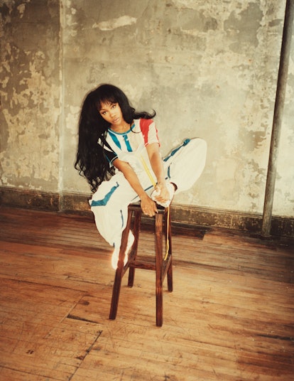 SZA sitting on a barstool, with both legs up, wearing a white pant suit with multicolored stripes