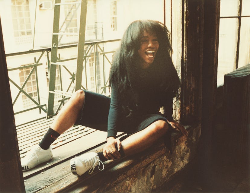 SZA sitting on a window sill laughing while wearing a black top and shorts