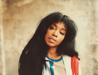 SZA tilting her head and looking straight at the camera in front of a brown wall
