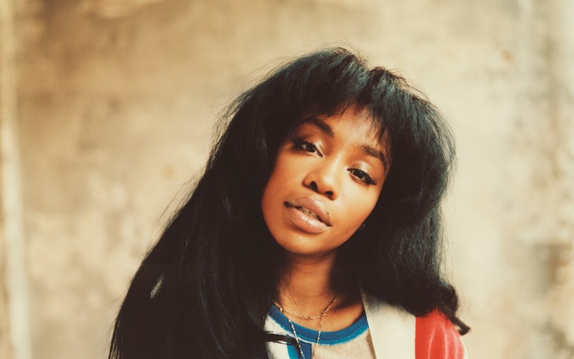 SZA tilting her head and looking straight at the camera in front of a brown wall