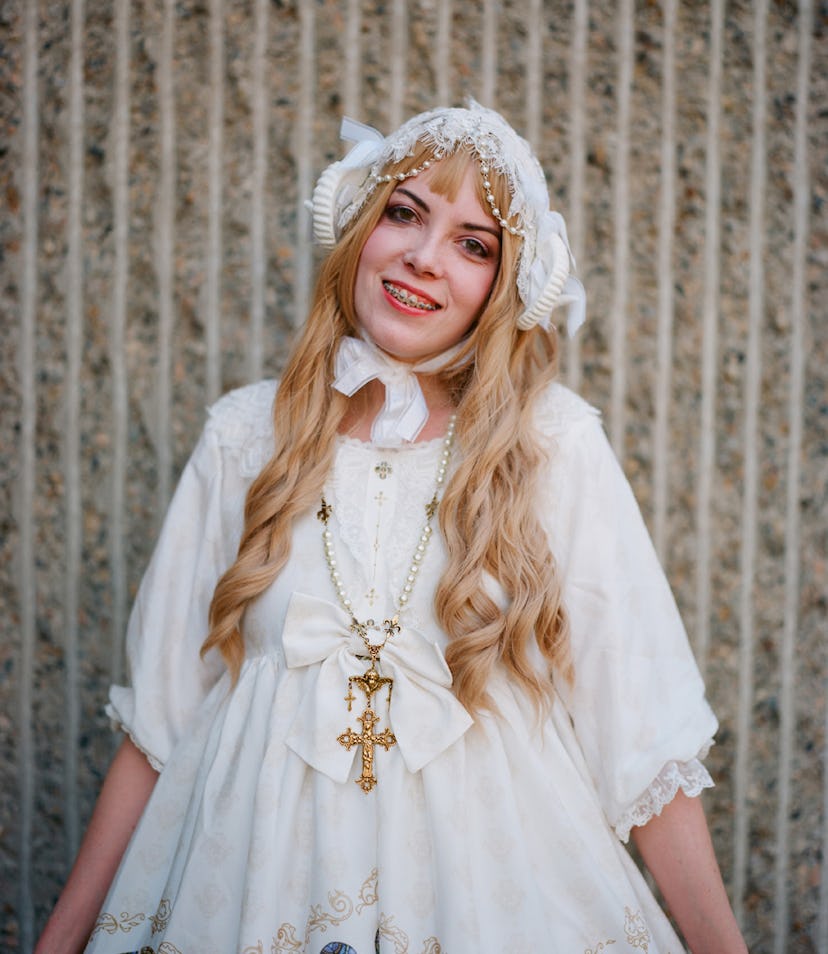 A blonde girl with braces posing while wearing a white dress and a white hat on her head 