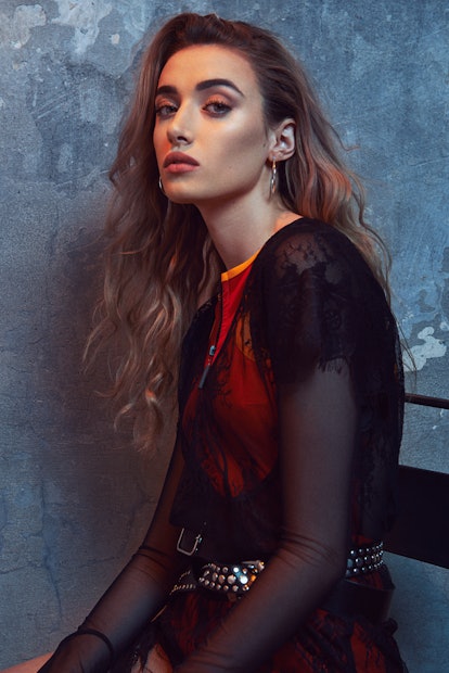 Olivia O’Brien leaning against a wall with her hair down while wearing a sheer black shirt and a red...