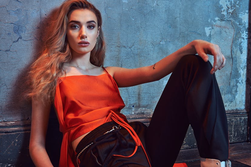 Olivia O’Brien leaning back with one leg raised up while wearing an orange top and black pants