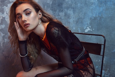 Olivia O’Brien posing in a chair while wearing a shirt with sheer sleeves and red detailing