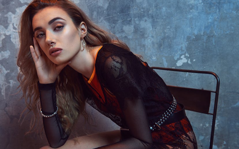 Olivia O’Brien posing in a chair while wearing a shirt with sheer sleeves and red detailing