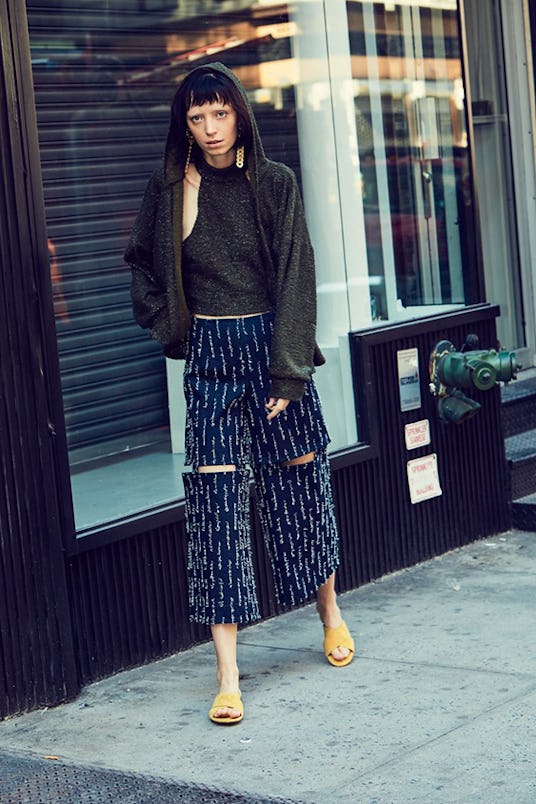 A model wearing navy blue, patterned pants, a black sleeveless shirt and hoodie and yellow sandals