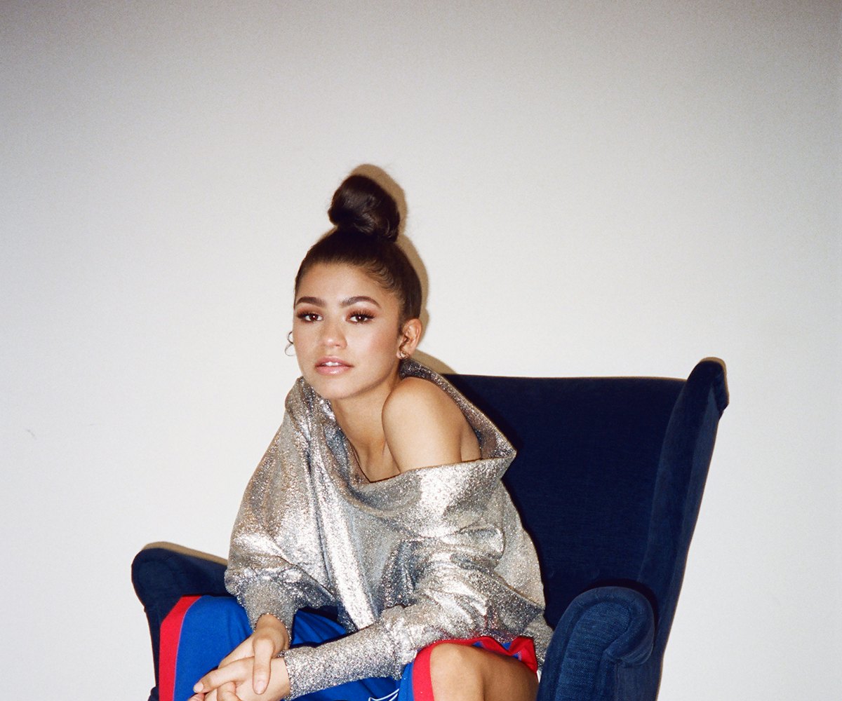 Zendaya sitting on an armchair while wearing a long blue skirt in combination with a shiny shirt 