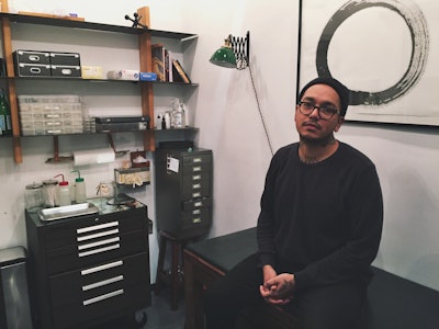 Adrian Castillo sitting in his piercing studio in a black shirt and a black beanie 